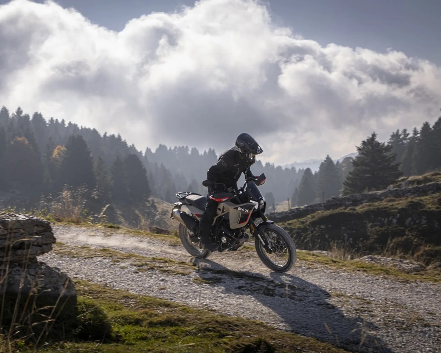 Motorcyclist in black gear riding a beige and black BCX dirt bike on a gravel path in a mountainous forest area.