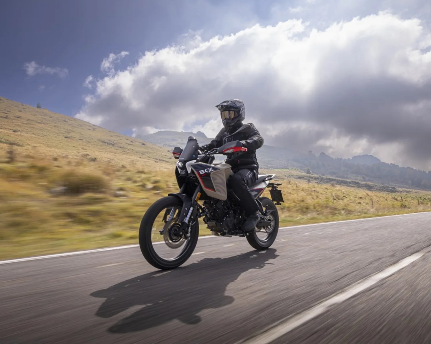 Person wearing black riding gear and helmet riding a black and white BCX motorcycle on a paved road with grassy hills and cloudy sky in the background.