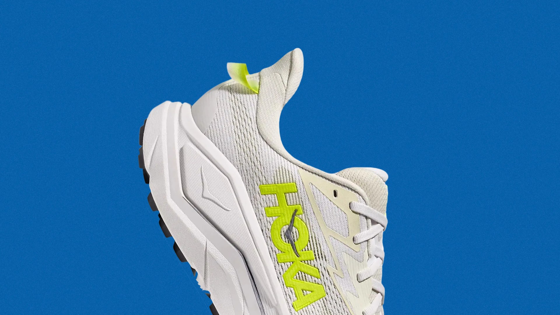 White HOKA running shoe with neon yellow logo and pull tab against a blue background.
