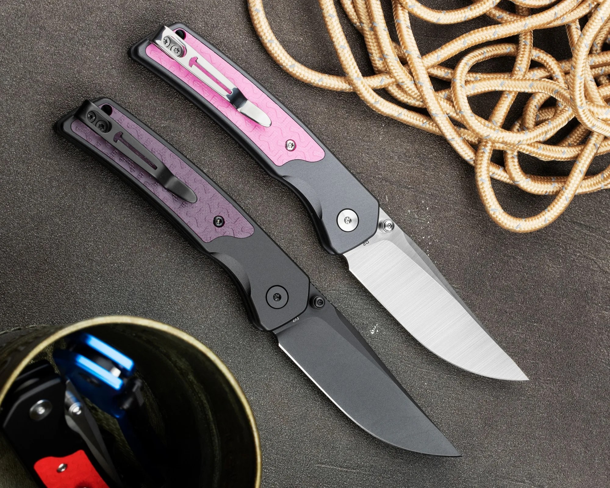 Two folding knives with black handles and purple and pink textured inlays, one with a silver blade and the other with a black blade, on a dark surface next to coiled rope.