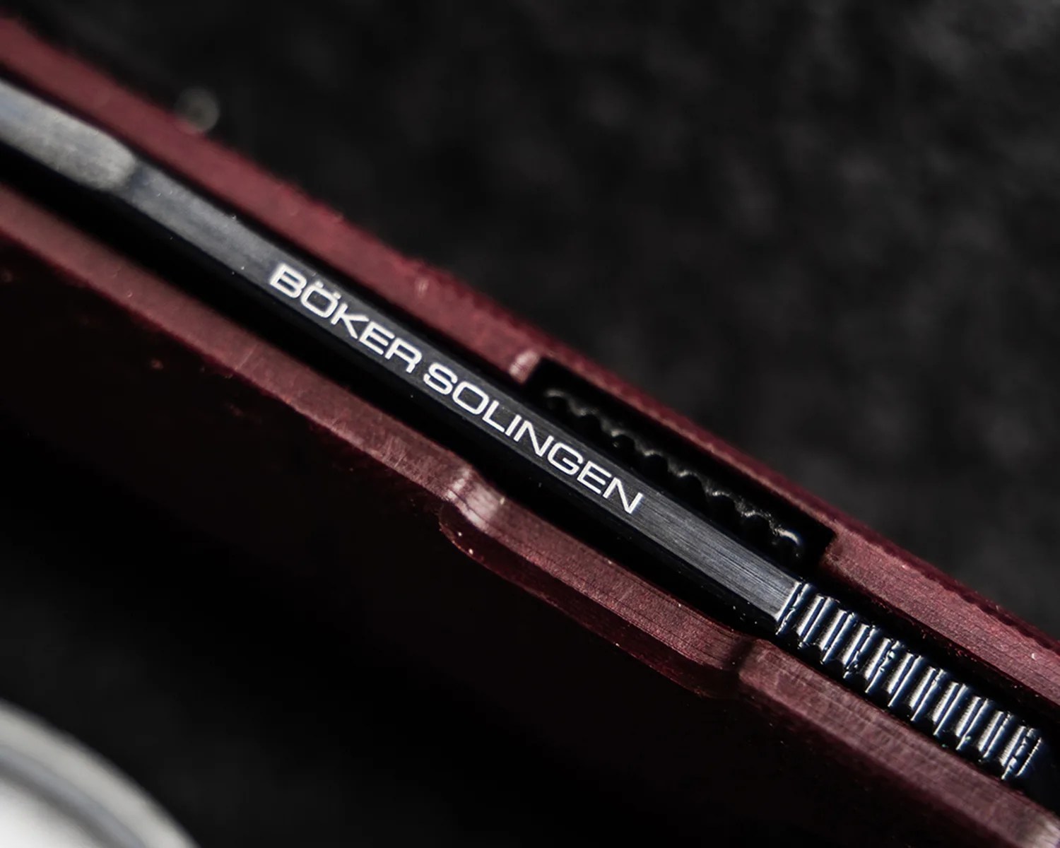 Close-up of a black metal knife spine with "BÖKER SOLINGEN" engraved, set in a dark red handle.