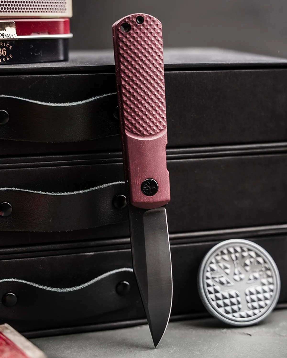 Folding knife with textured maroon handle and black blade standing upright against a black leather case.