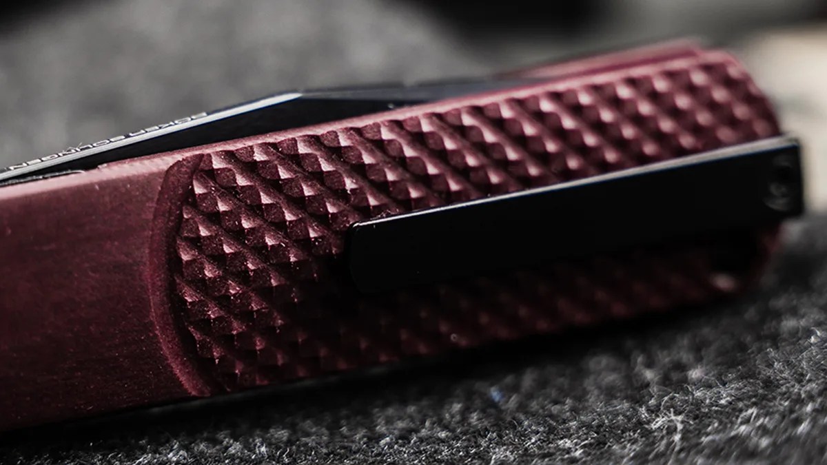 Close-up of a textured dark red folding knife handle with a black pocket clip on a gray fabric surface.