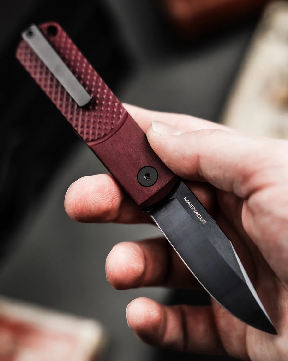 Hand holding a folding knife with a textured maroon handle and a black blade labeled "MAGNACUT.