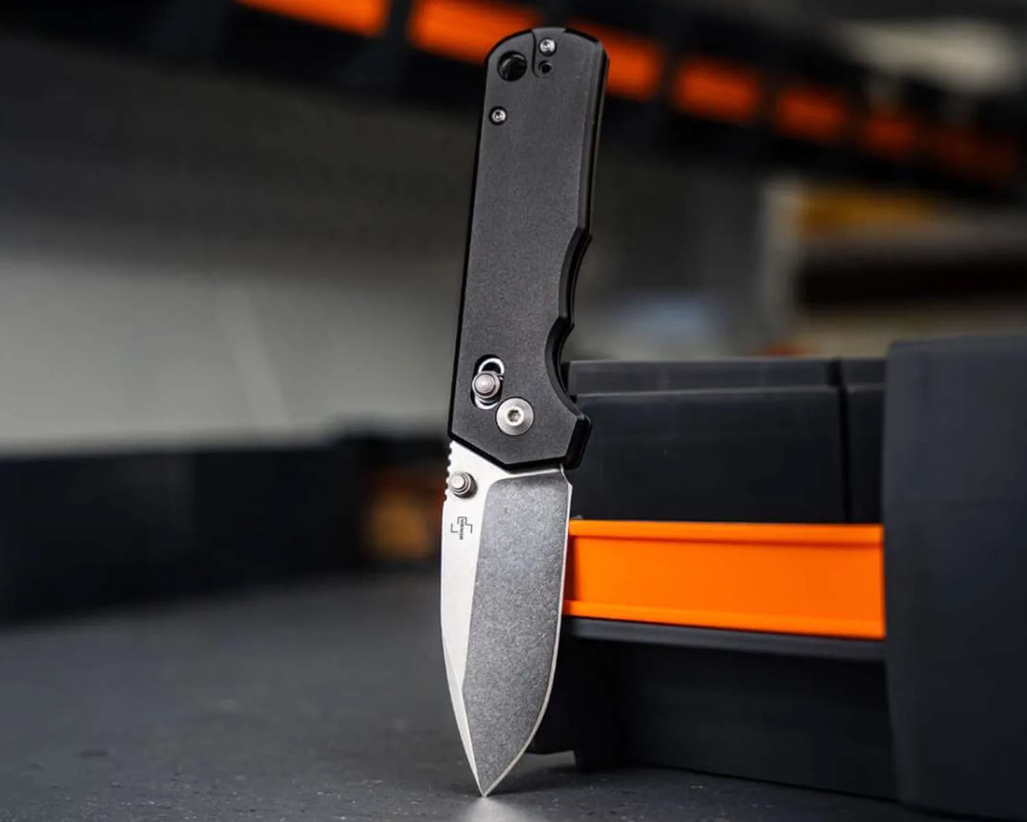 Folding knife with a stonewashed blade and black textured handle standing upright on a dark surface next to an orange and black box.