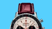 Close-up of a Breitling watch face with rose gold hands, black subdials, and a brown leather strap against a blue background.