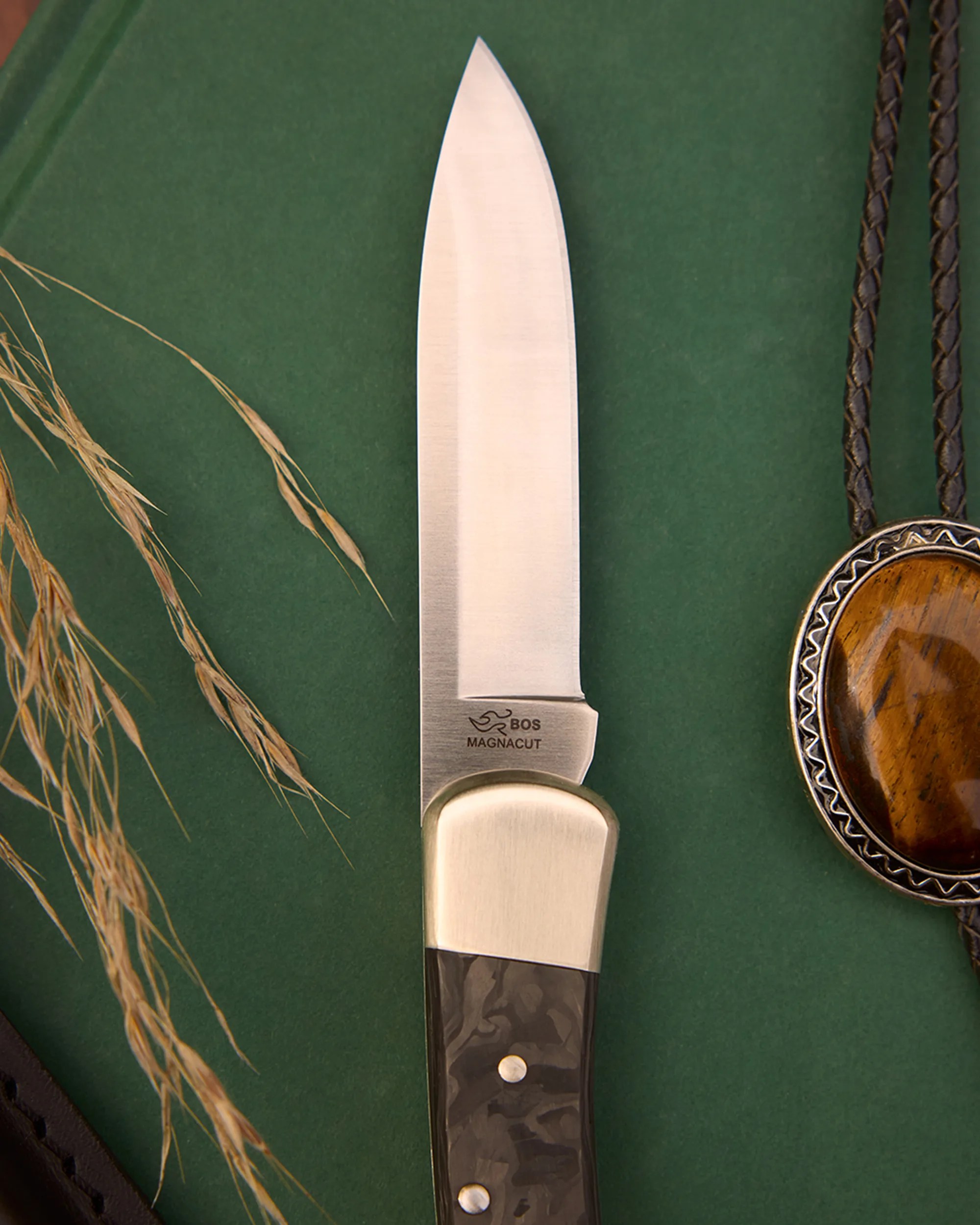 Knife with a polished steel blade marked "BOS Magnacut" and a handle with a brass bolster and dark patterned grip on a green surface.