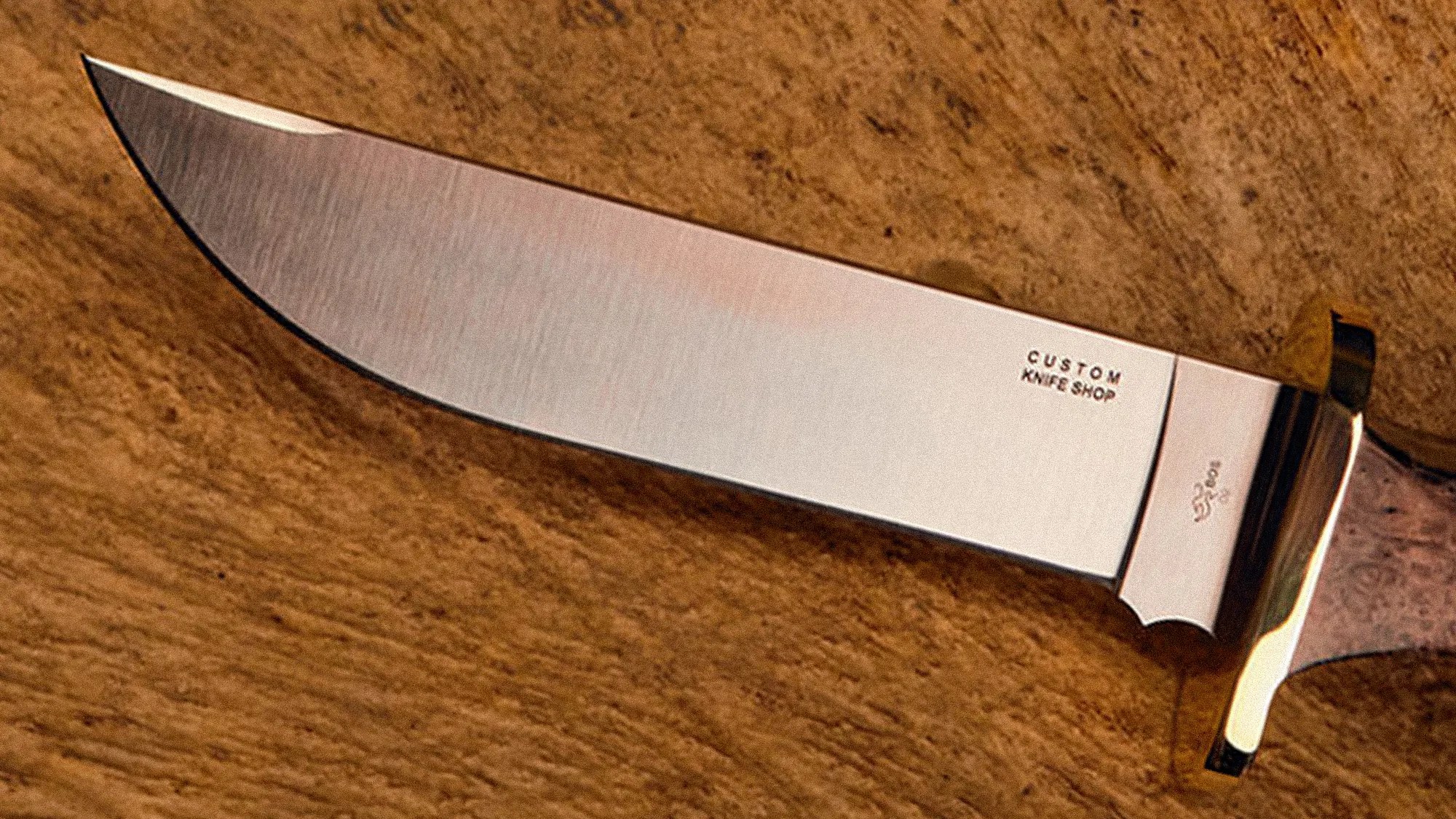 Knife blade with "CUSTOM KNIFE SHOP" engraving and a polished guard on a wooden surface.
