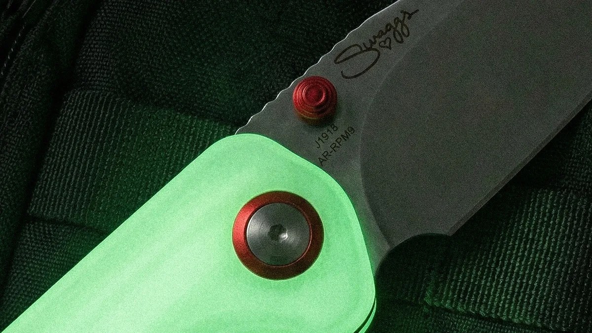 Close-up of a folding knife with a glowing green handle, red pivot, and a stainless steel blade engraved with "J1918 AR-RPM9" and "Sugggs.