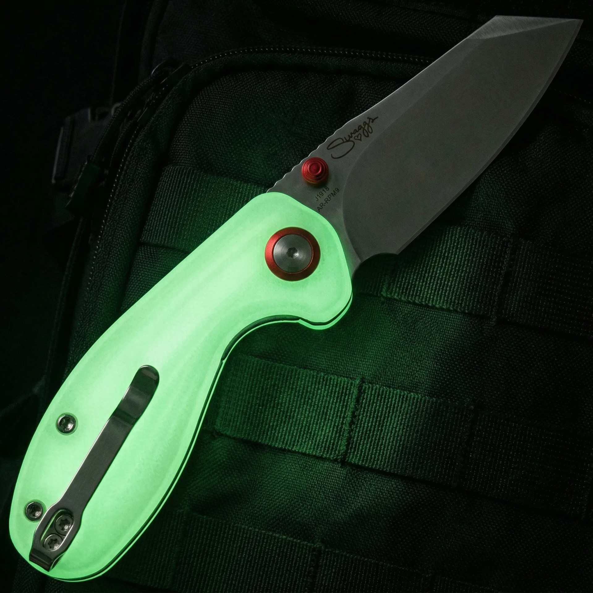 Folding knife with a glowing green handle, red accents, and a matte silver blade resting on black fabric.