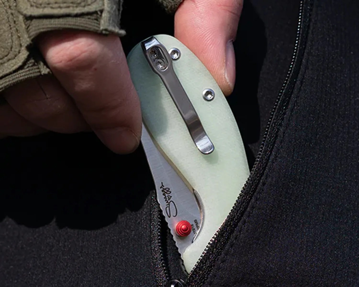 Folding pocket knife with light green handle and red thumb stud clipped inside a black zippered pocket.