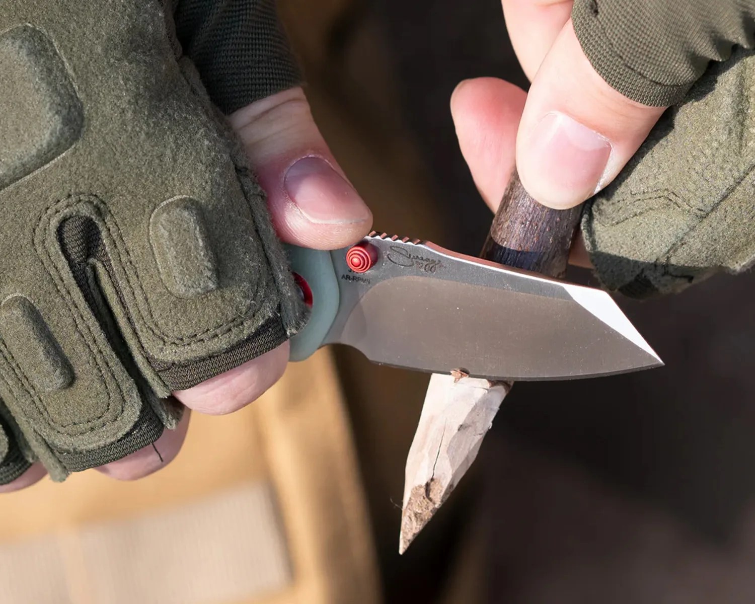 Close-up of a person wearing green tactical gloves carving a pointed wooden stick with a fixed-blade knife with a wooden handle.