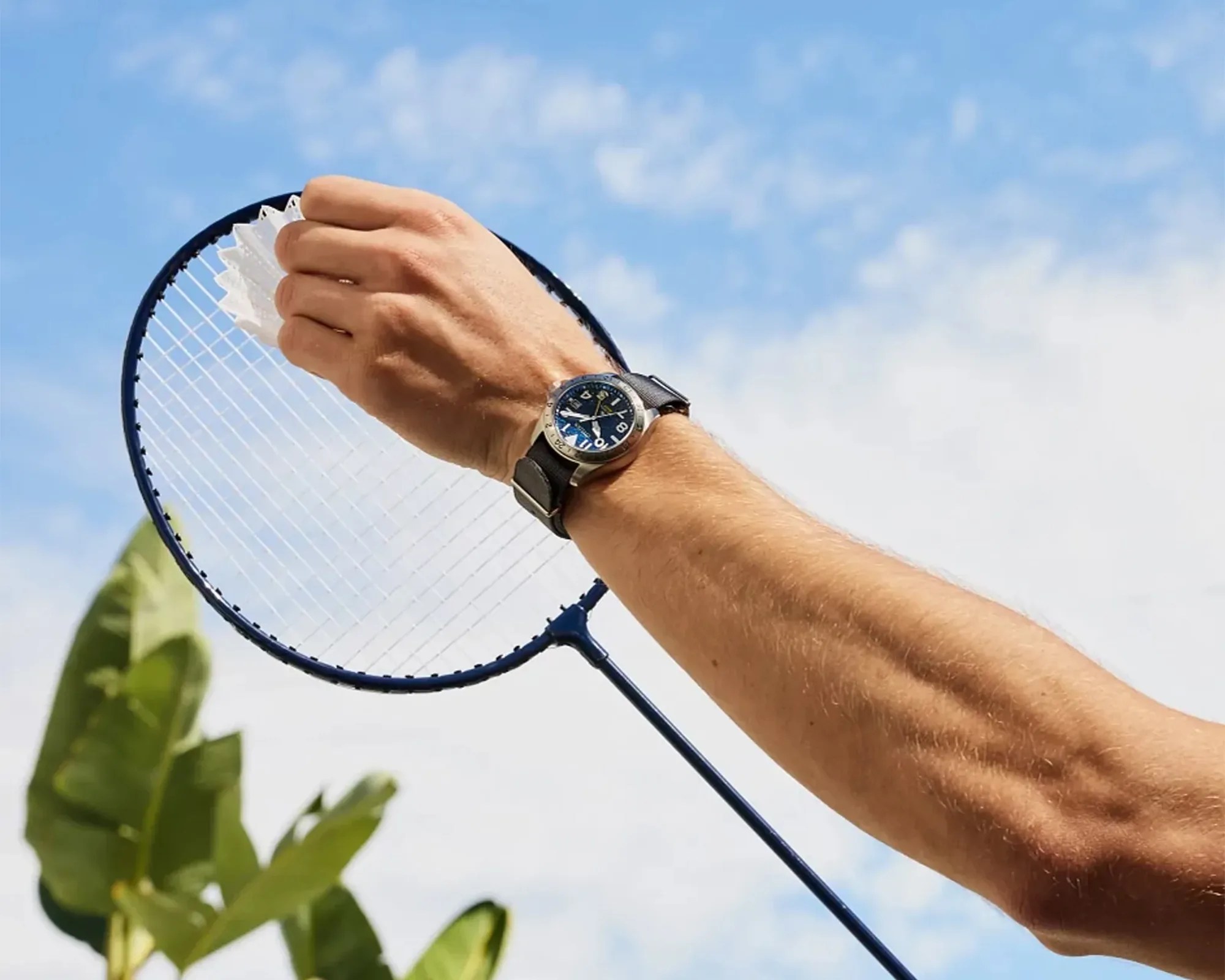 Arm wearing a black wristwatch holding a white shuttlecock over a blue badminton racket against a sky background.