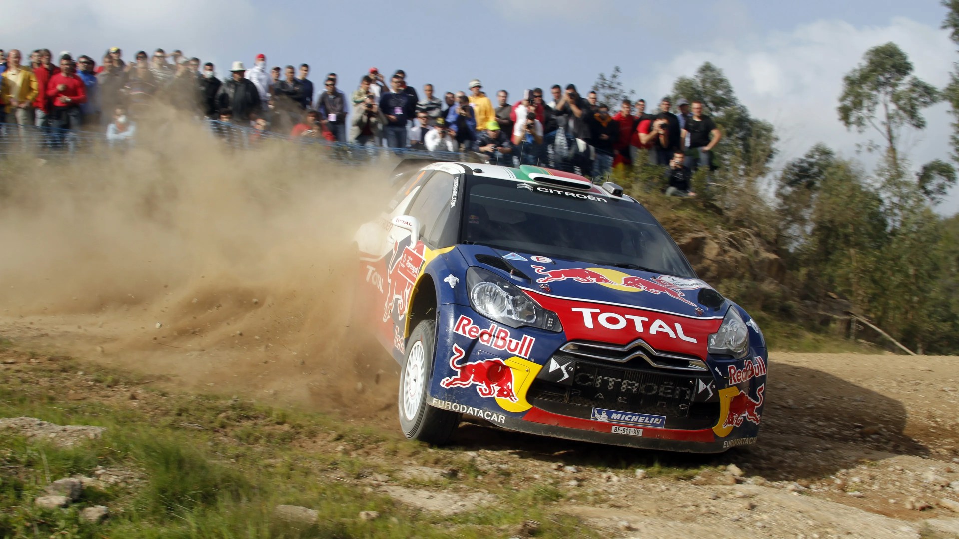 Citroën rally car with Red Bull and Total logos kicking up dust on a dirt track with spectators behind.