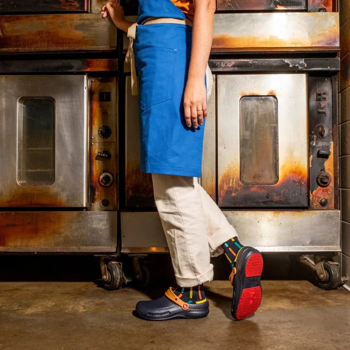 Person wearing a blue apron, beige pants, colorful striped socks, and black slip-resistant clogs with red soles in a kitchen.
