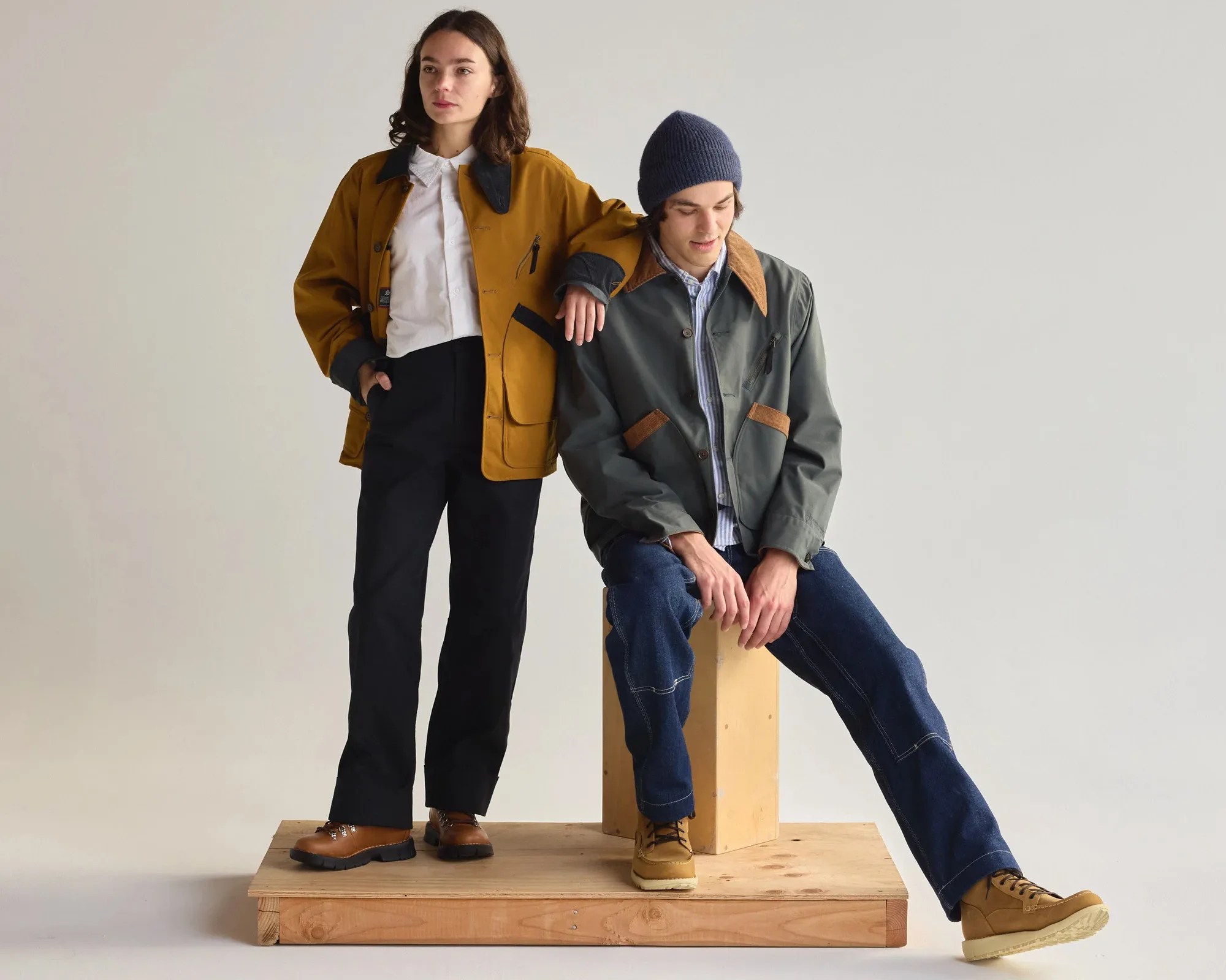Two people wearing casual jackets and boots, one standing in a mustard jacket and black pants, the other sitting in a green jacket, blue jeans, and a blue beanie.