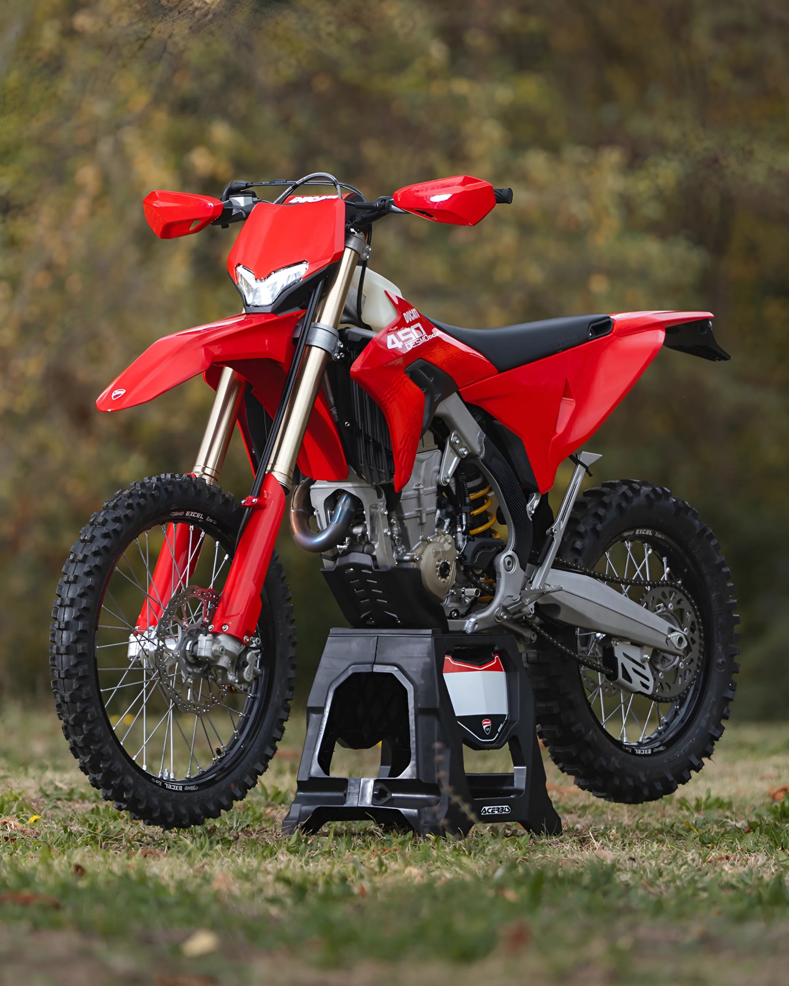 Red Ducati 450 motocross bike on a black stand on grass with blurred trees in the background.
