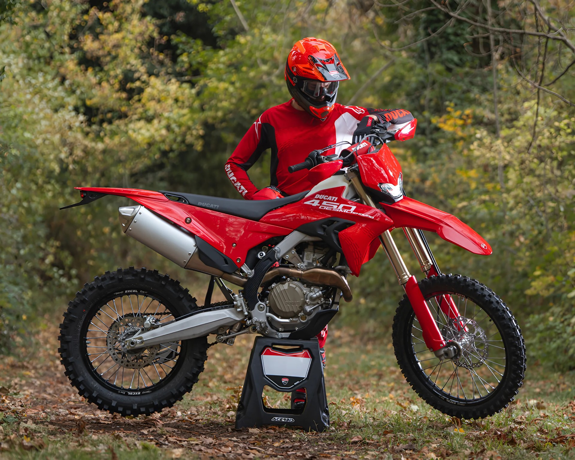 Red Ducati 450 dirt bike on a black stand with a rider in red gear and helmet in a forest setting.