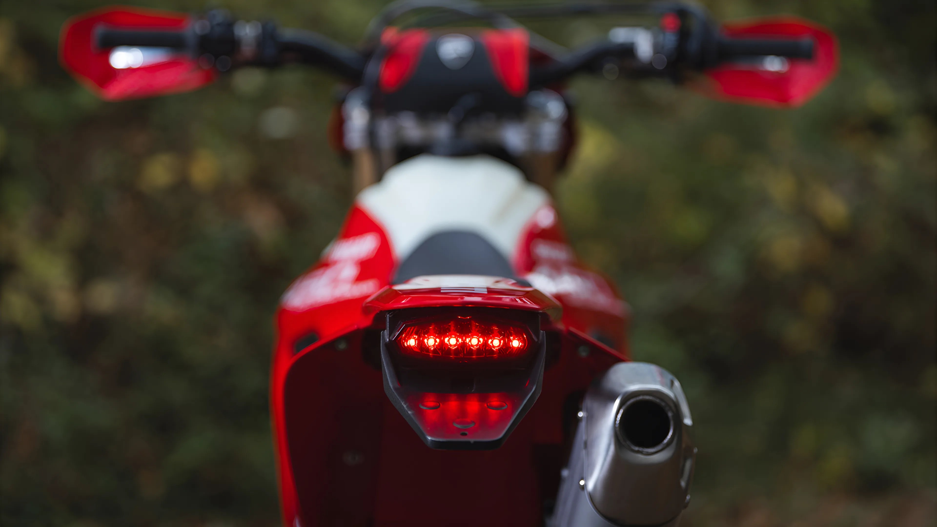 Rear view of a red motorcycle with illuminated tail light and silver exhaust pipe against a blurred natural background.