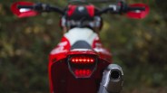 Rear view of a red motorcycle with illuminated tail light and silver exhaust pipe against a blurred natural background.