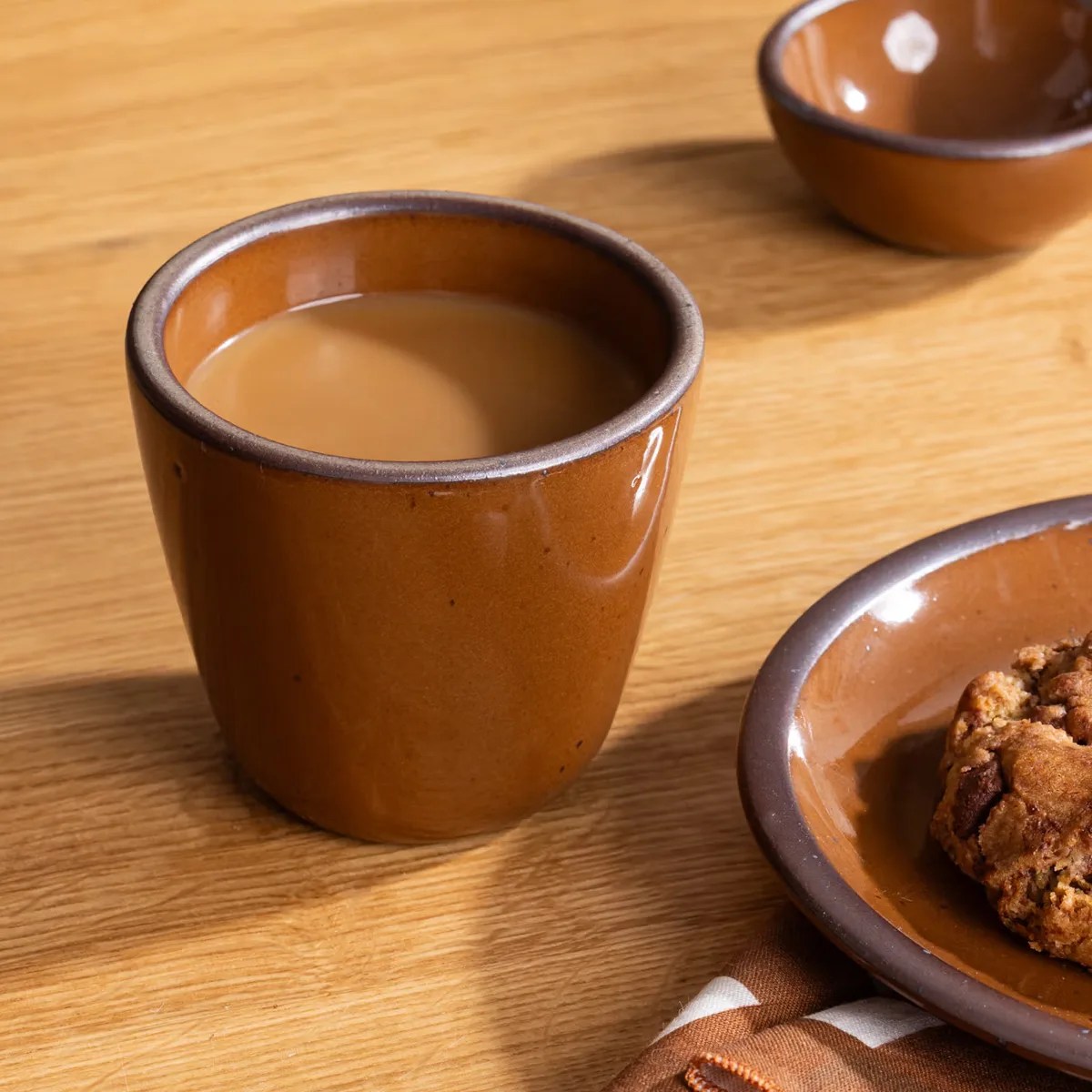 Brown ceramic cup filled with coffee on a wooden table next to a matching plate with a cookie.