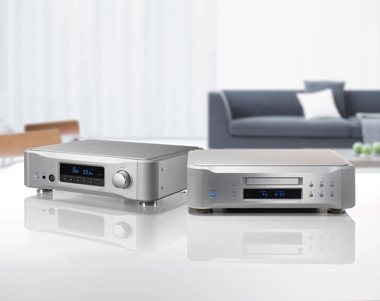 Two silver audio components with digital displays on a white reflective surface in a modern living room setting.
