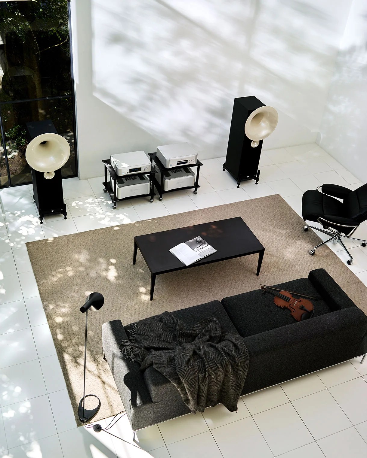 Modern living room with black sofa, beige rug, black coffee table with open book, black floor lamp, violin on sofa, and large horn speakers.
