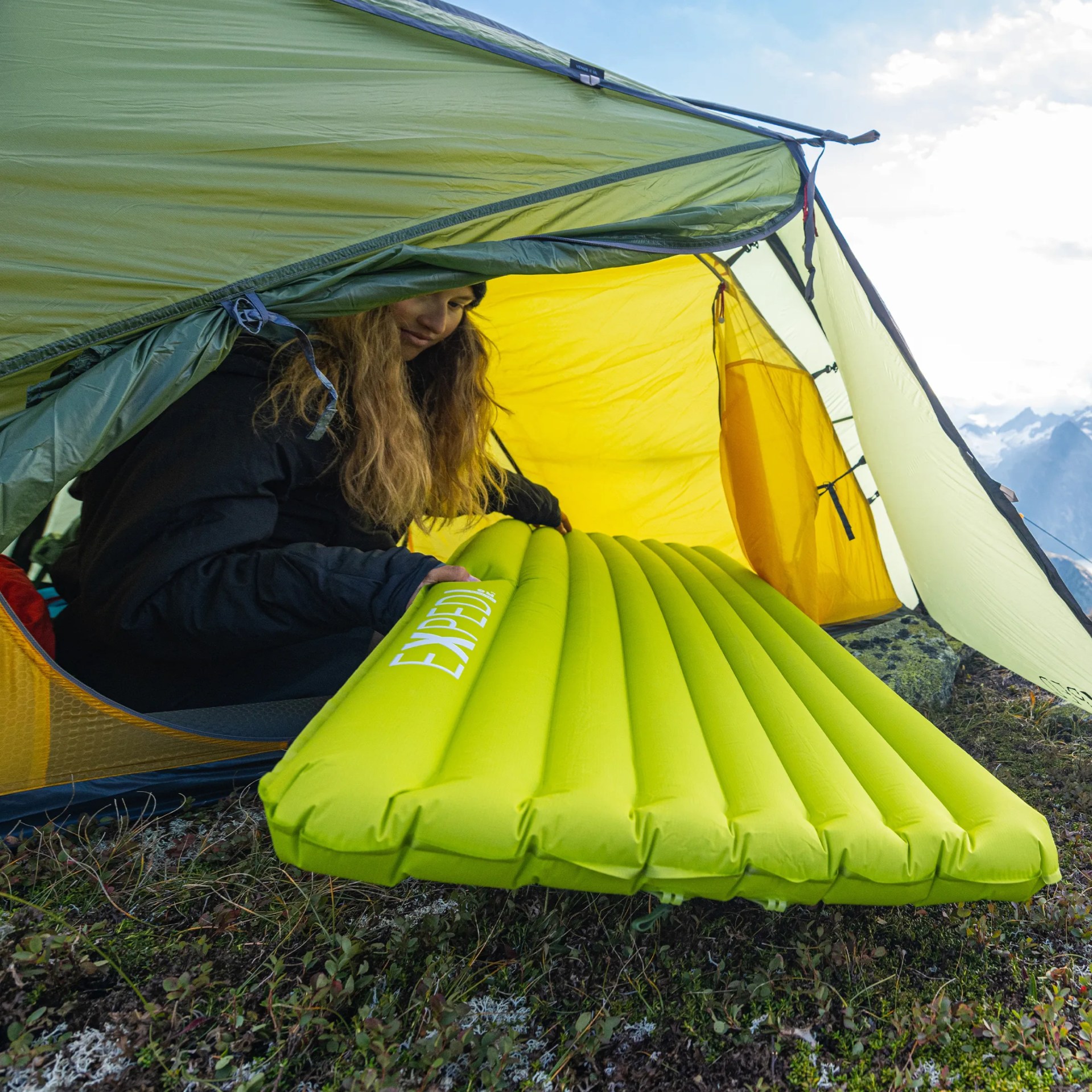 Person inside a green and yellow tent holding a bright green inflatable sleeping pad labeled "EXPED" on grassy ground.