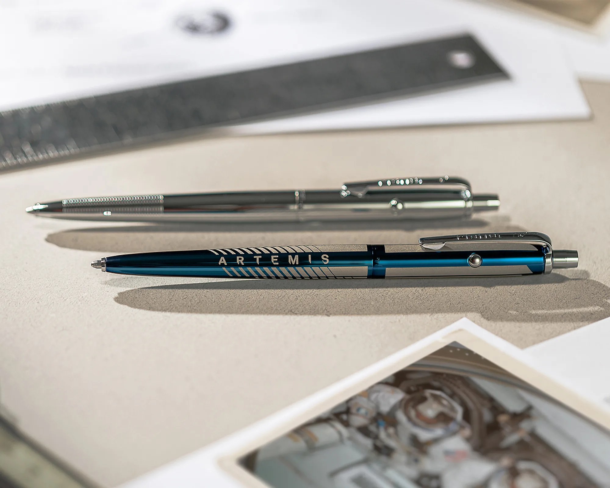 Two metallic pens, one silver and one blue with "ARTEMIS" text, lying on a beige surface with a ruler and photo nearby.