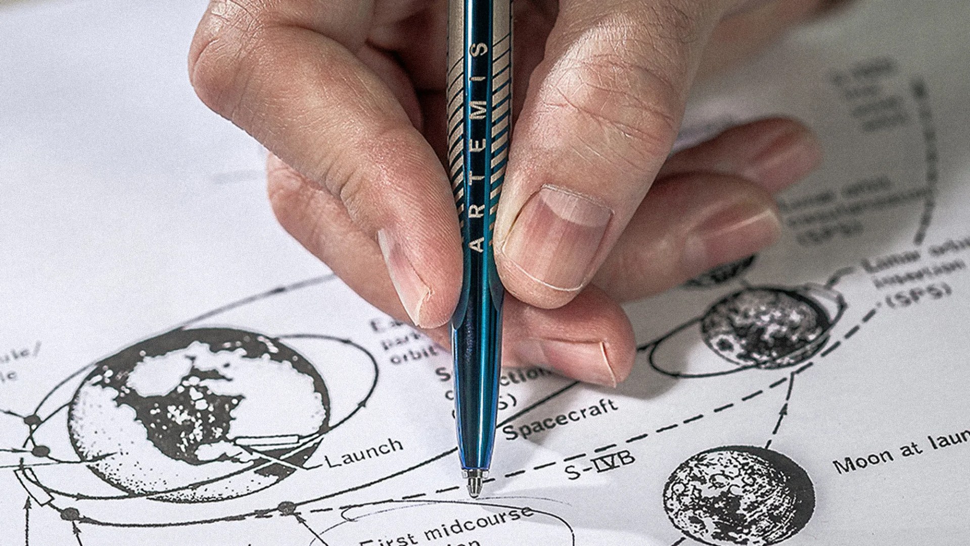 Hand holding a blue pen labeled "ARTEMIS" pointing at a diagram of Earth, Moon, and spacecraft trajectories.