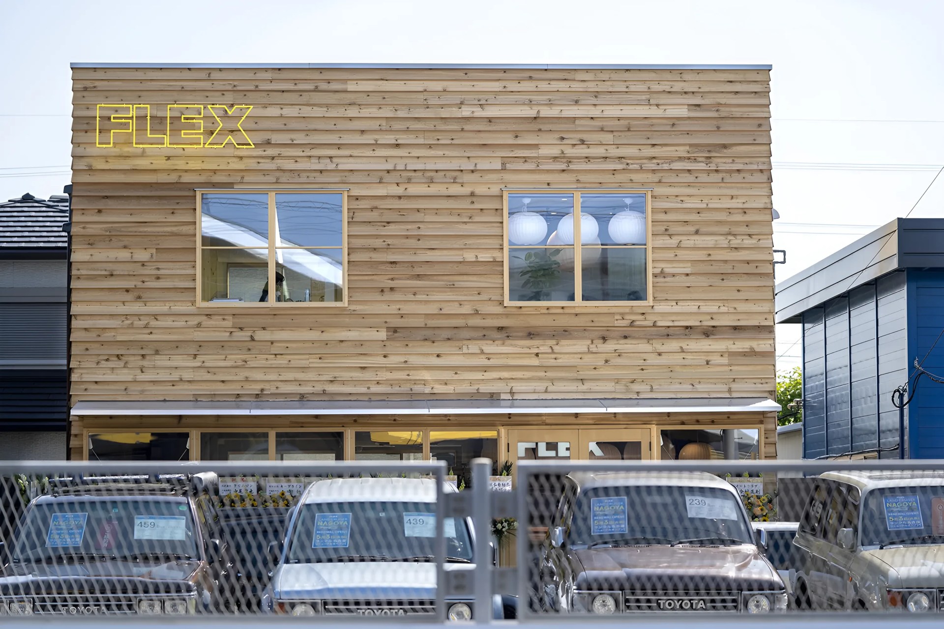 Wood-paneled two-story building with yellow "FLEX" sign and parked Toyota vehicles behind a chain-link fence.
