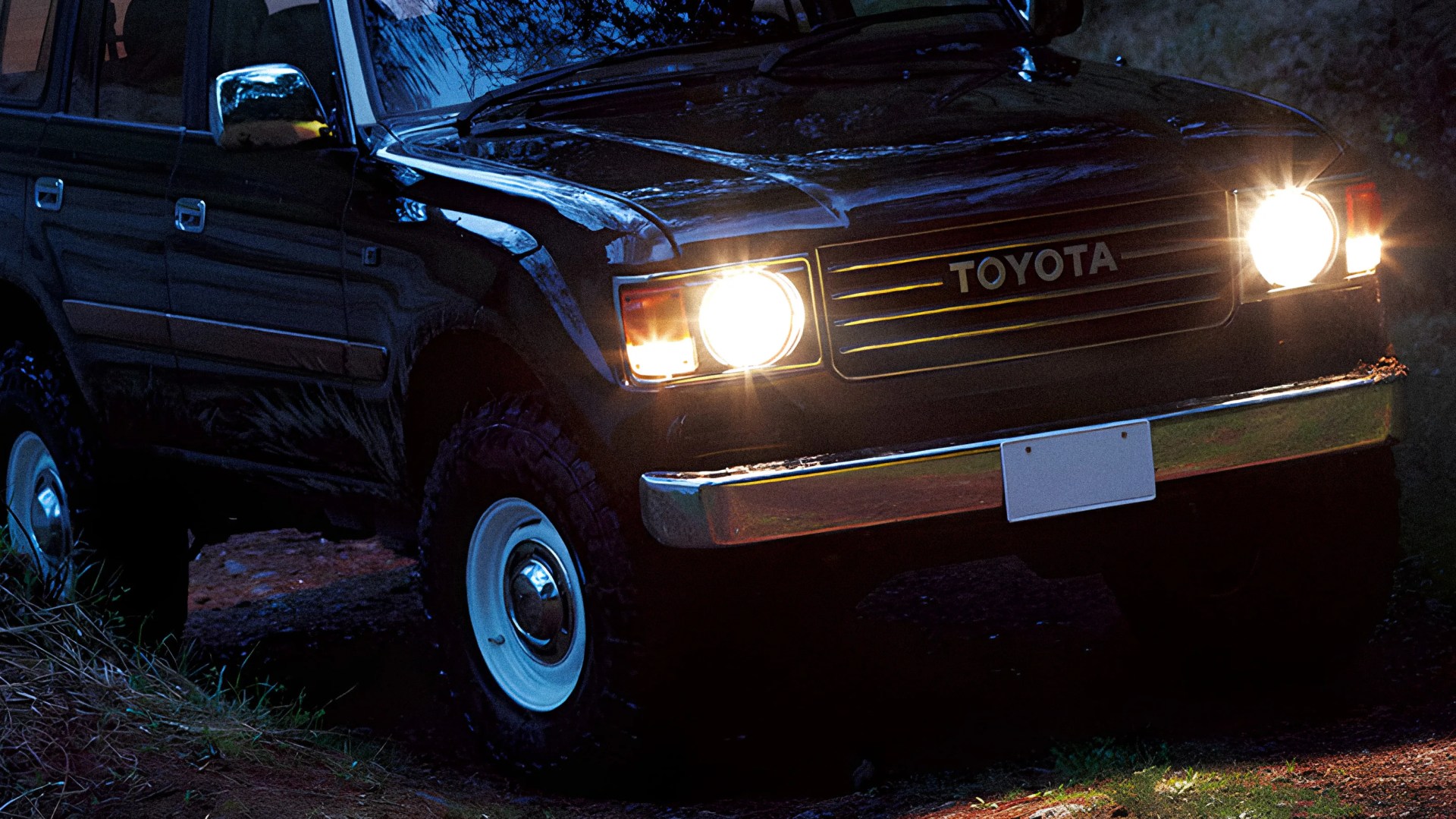 Black Toyota SUV with headlights on, parked on a dirt path at night.