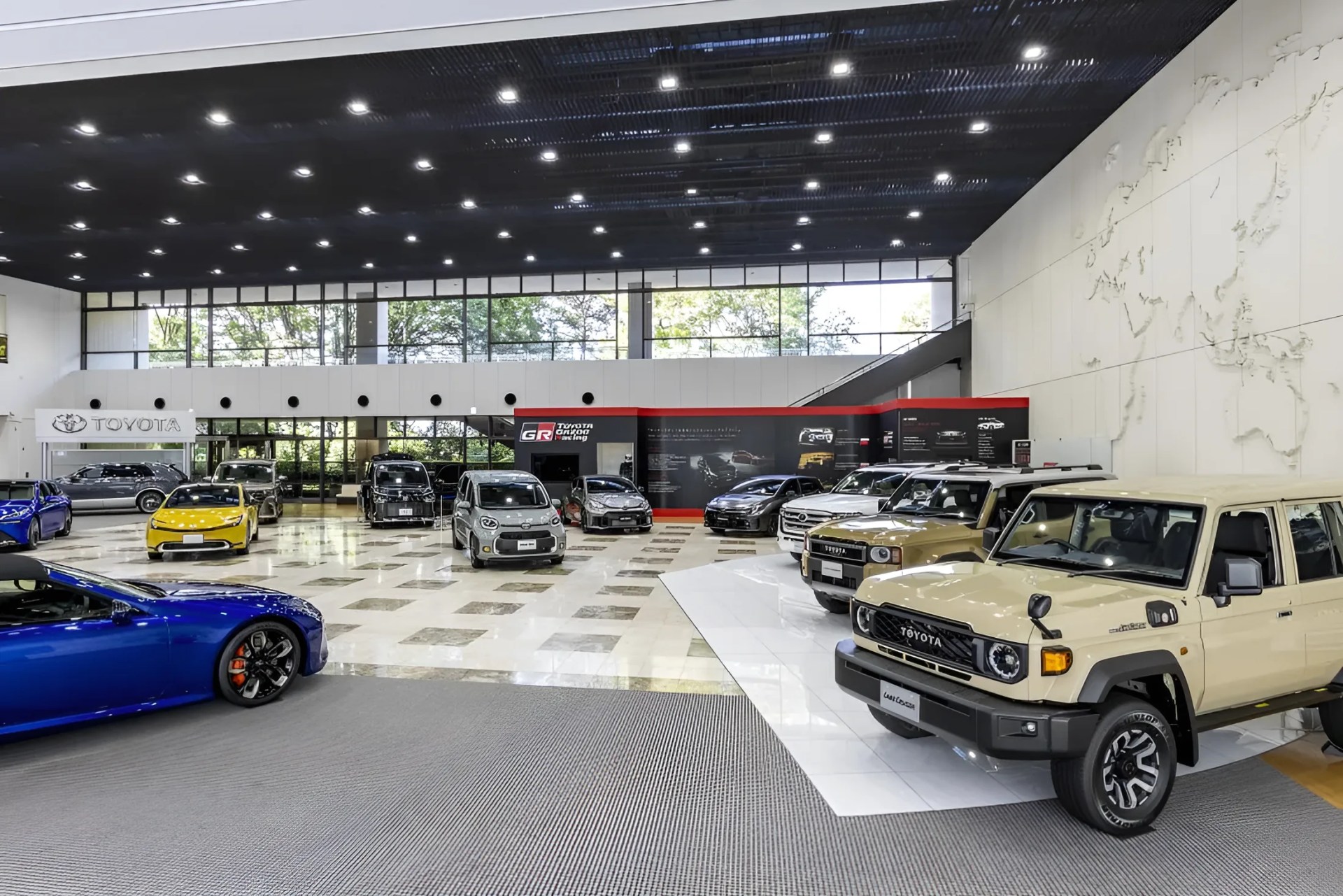 Showroom with various Toyota cars including beige Land Cruiser SUVs, a yellow compact car, and a blue sports car under bright ceiling lights.