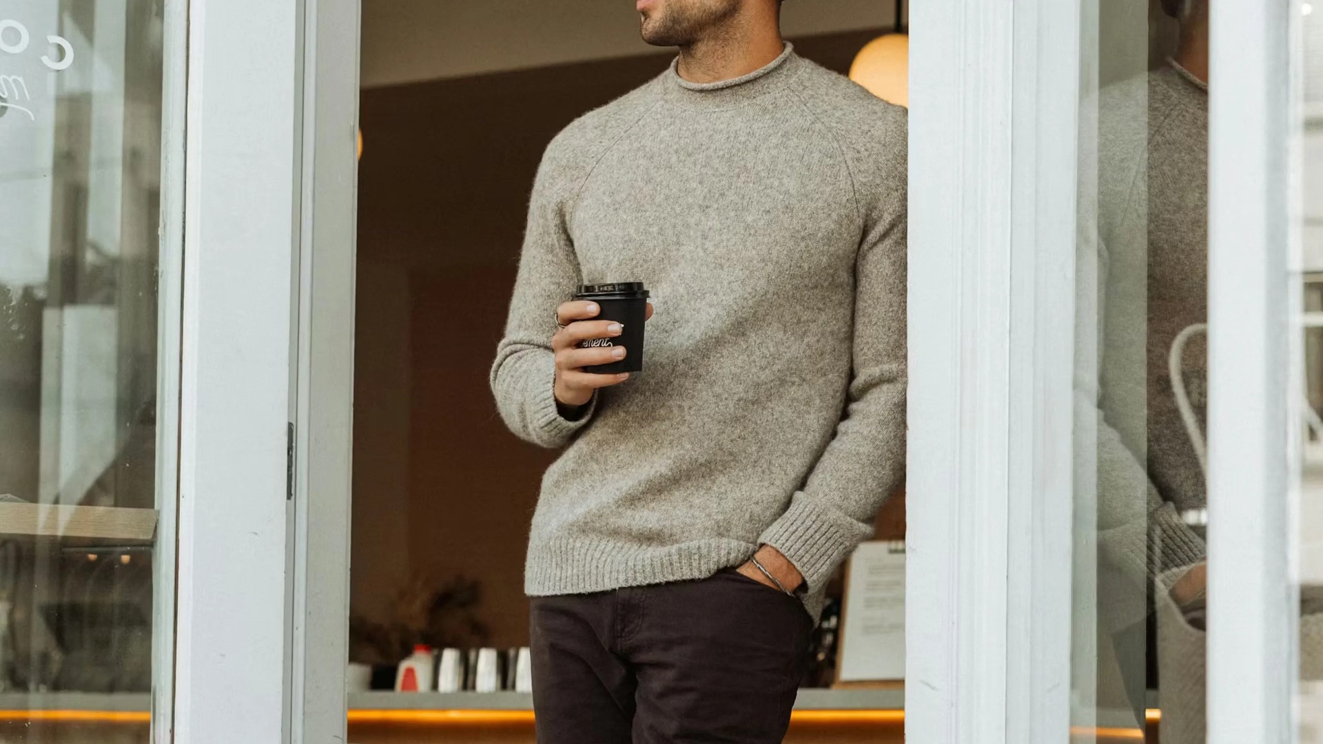 Man wearing a light gray wool sweater holding a black coffee cup standing by a glass door.