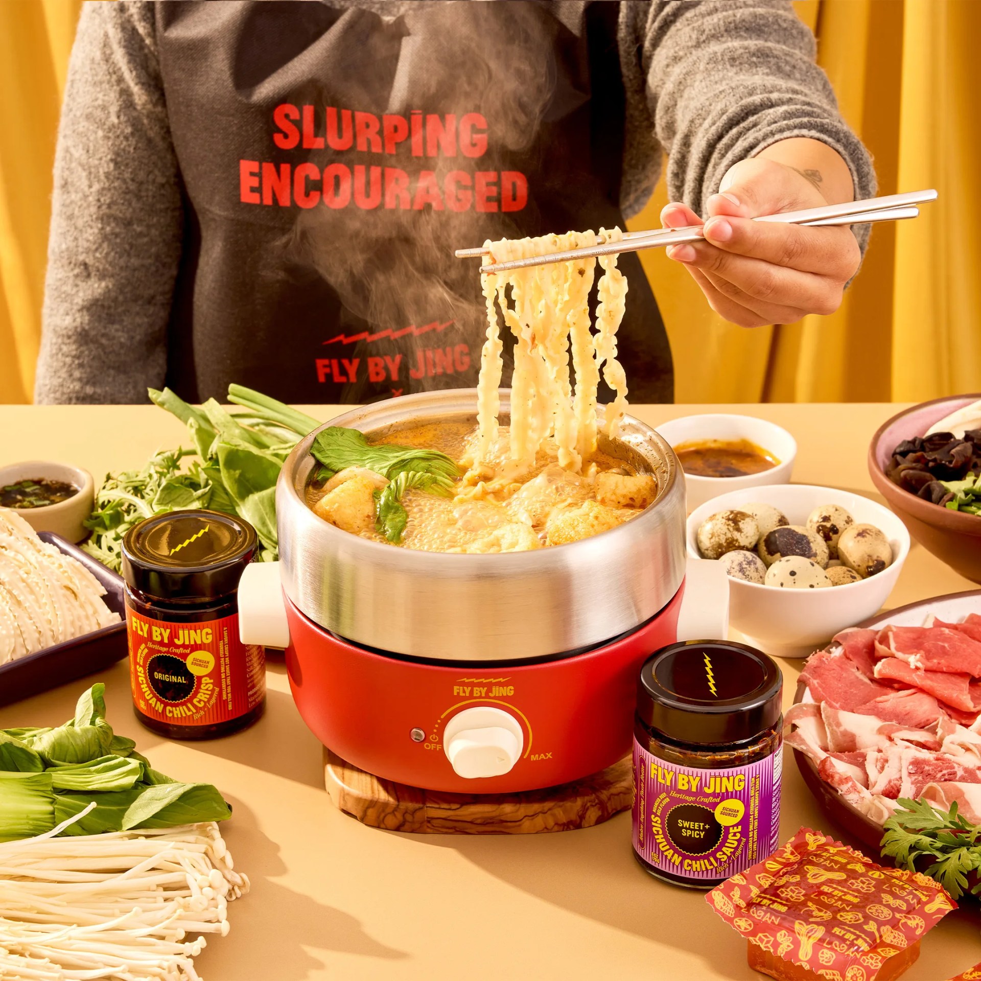 Red electric hot pot with steaming noodles held by chopsticks, surrounded by fresh greens, quail eggs, sliced meat, and Fly By Jing chili sauce jars.