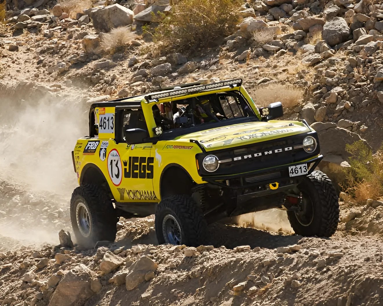 Yellow off-road Ford Bronco with large tires and racing decals driving on rocky terrain.