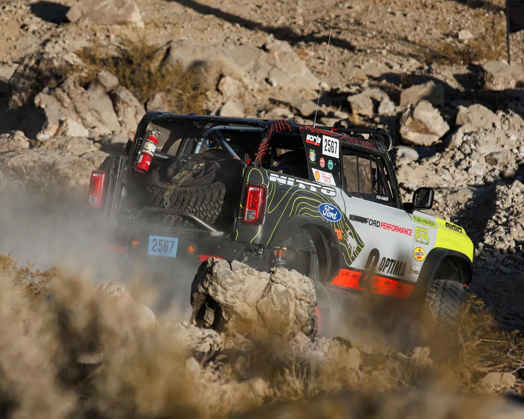 Off-road Ford Bronco with racing decals and spare tire driving on rocky terrain.