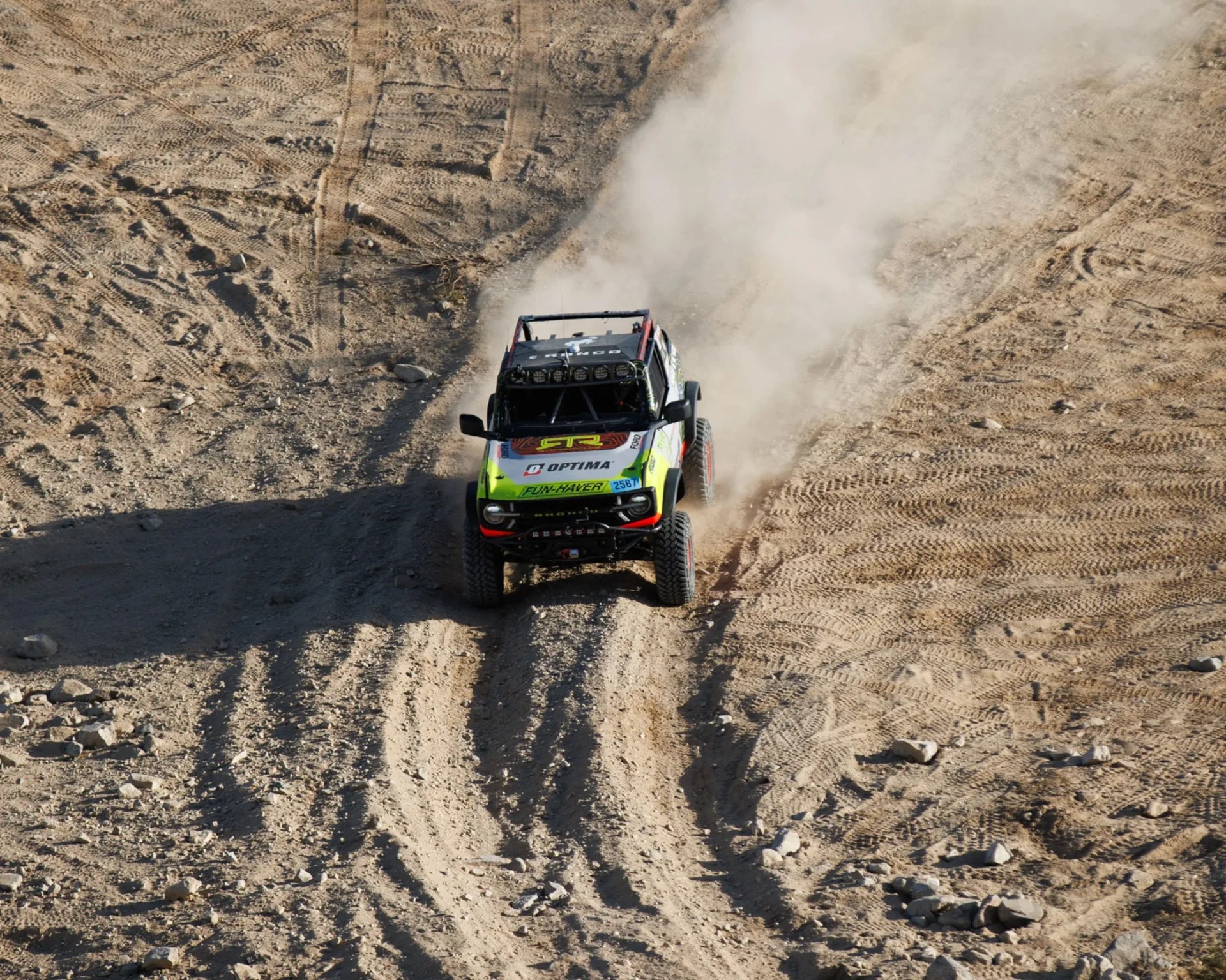 Off-road racing vehicle kicking up dust on a rugged dirt track with tire marks and scattered rocks.