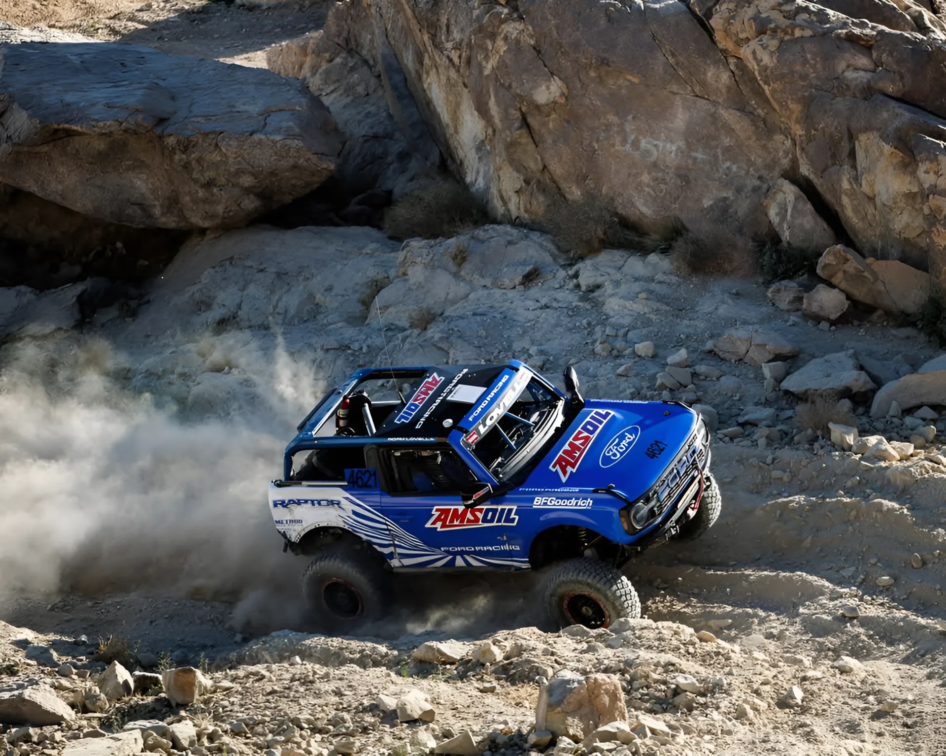 Blue and white off-road racing truck with AMSOIL and Ford logos kicking up dust on rocky terrain.
