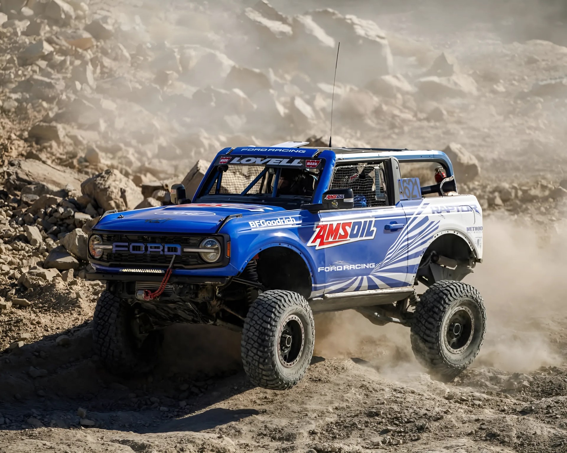 Blue Ford off-road racing truck with large tires kicking up dust on rocky terrain.