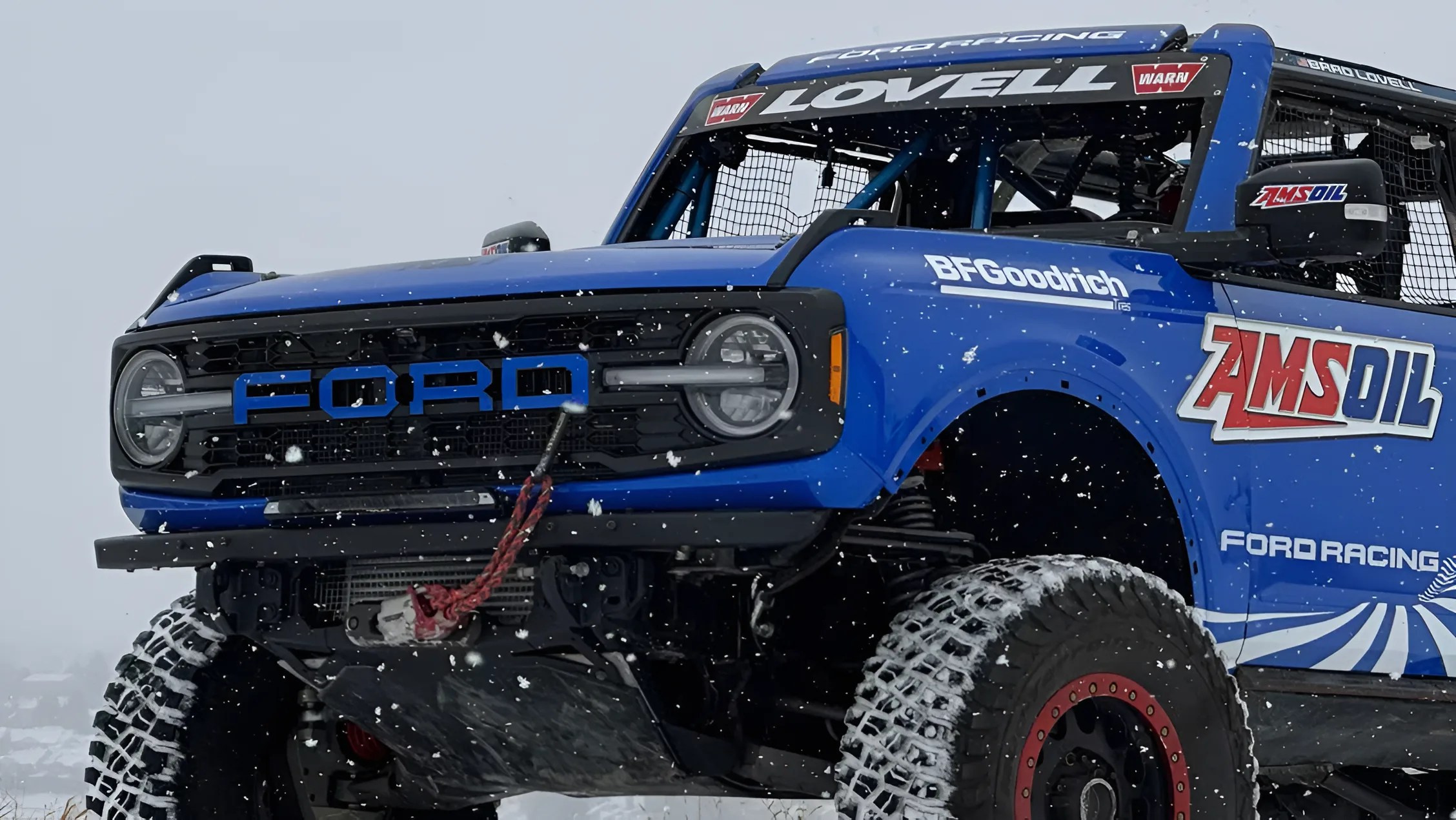 Blue Ford off-road racing truck with large BFGoodrich tires and AMSOIL branding in falling snow.