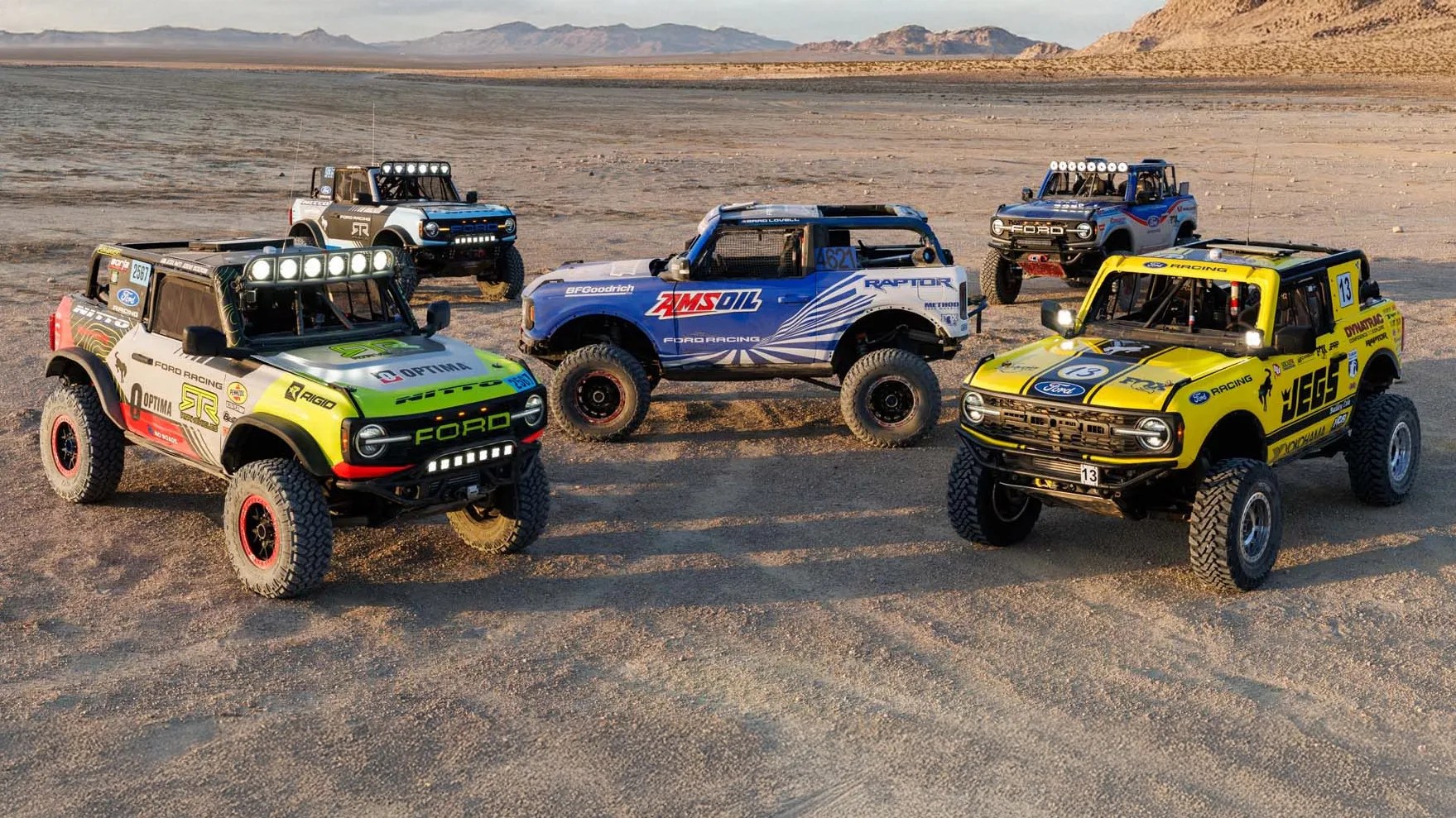 Five off-road Ford Broncos with racing decals and large off-road tires parked on a desert terrain with mountains in the background.