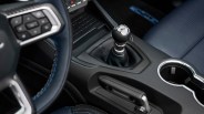 Black leather manual gear shift knob with chrome accents in a car interior with blue stitching and cup holder.