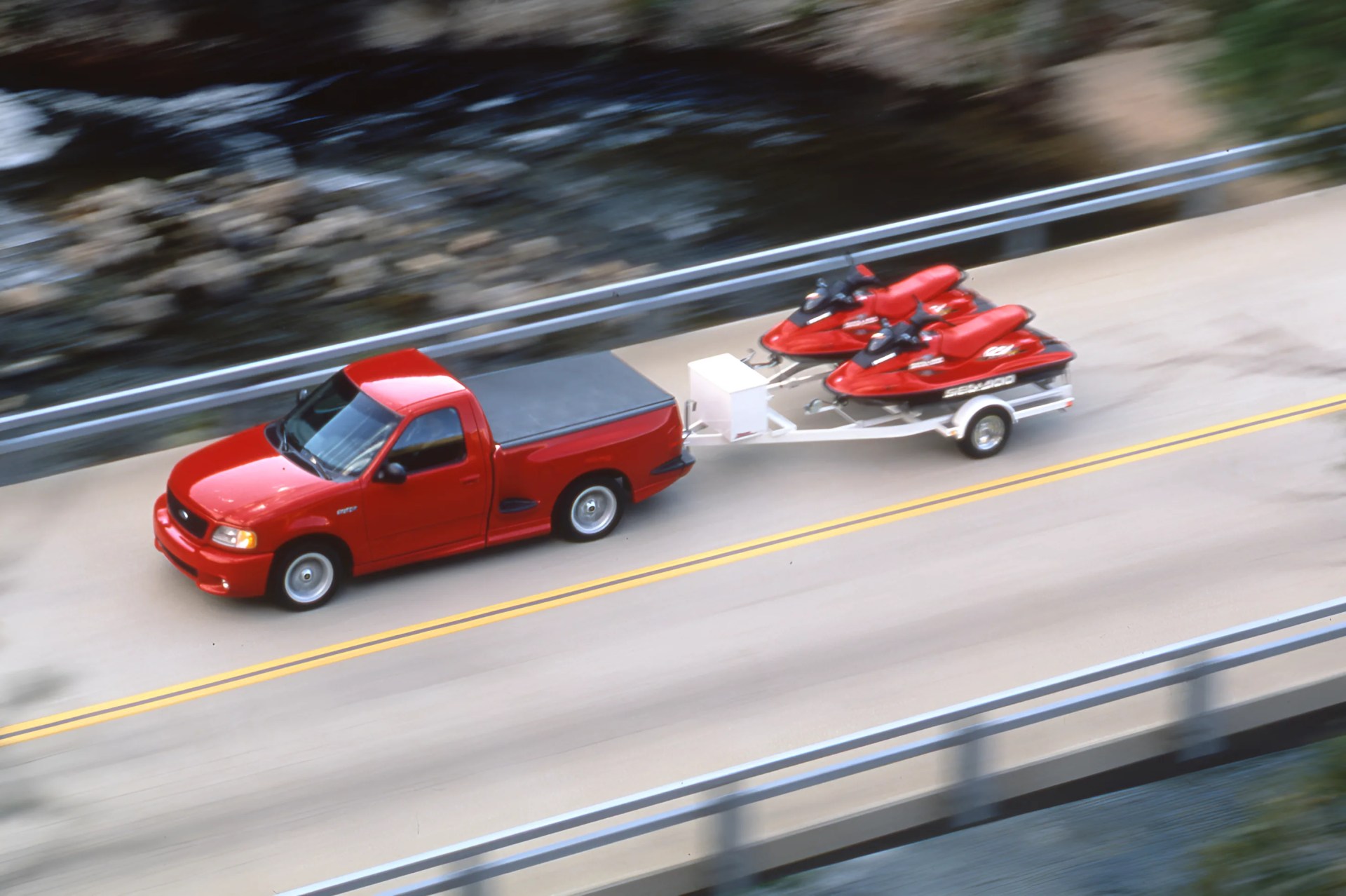 Red pickup truck towing a trailer with two red Sea-Doo jet skis on a road beside a river.