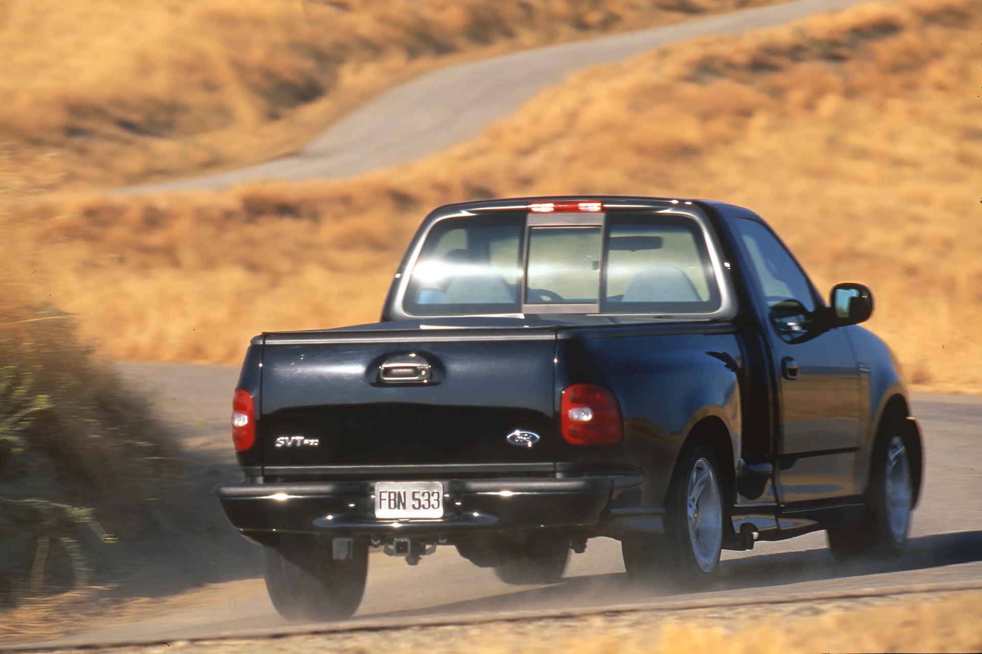 Black Ford SVT Lightning pickup truck driving on a winding road with dry grass hills in the background.