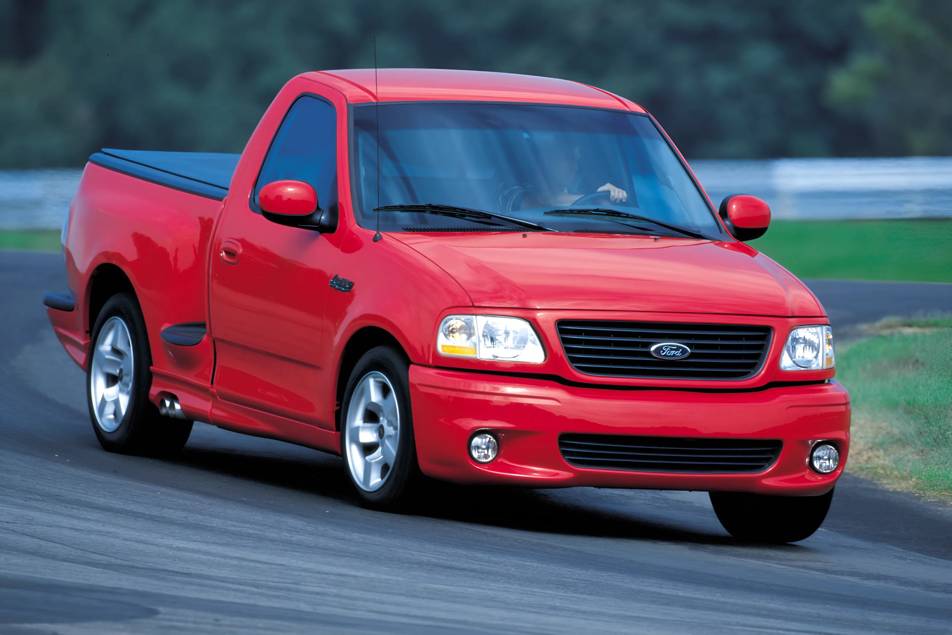 Red Ford pickup truck with covered bed driving on a paved road.
