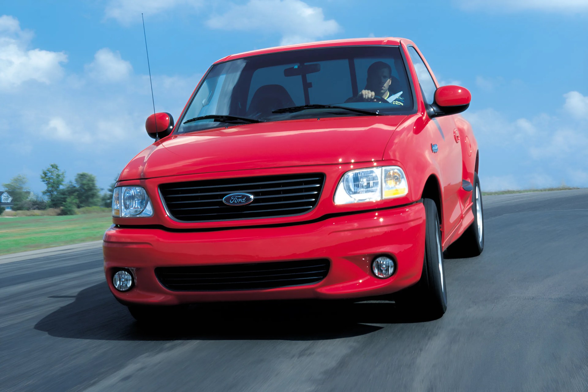 Red Ford pickup truck driving on a curved road under a blue sky.