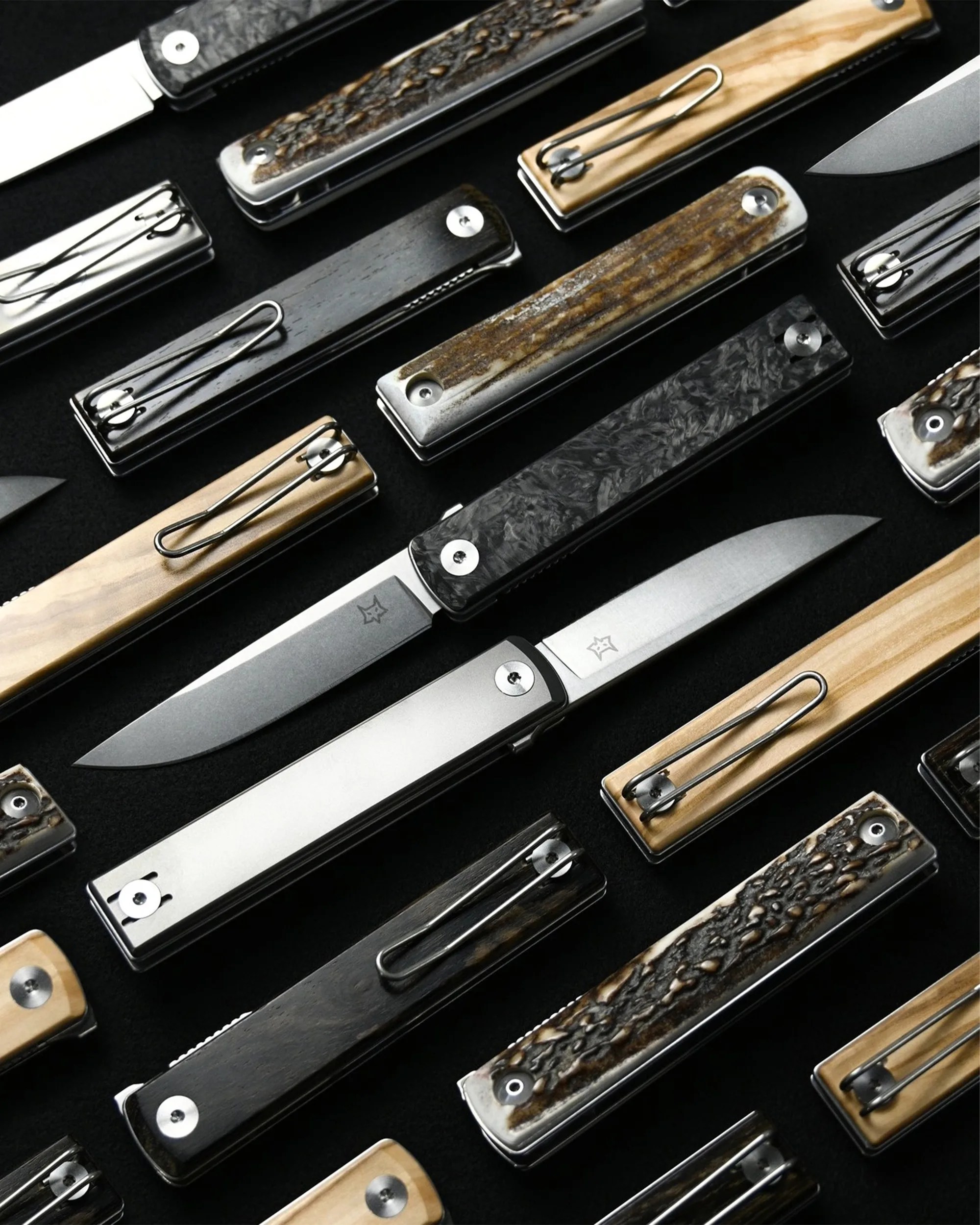 Multiple folding pocket knives with various textured and wooden handles arranged on a black surface.