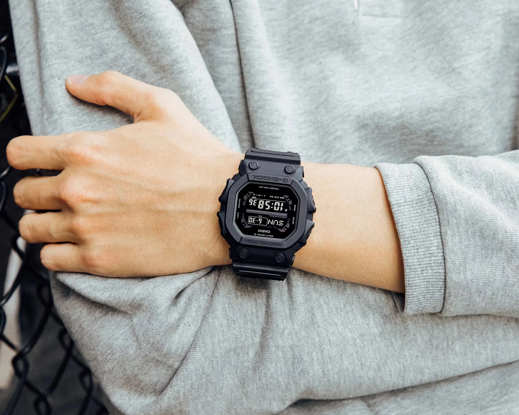 Black Casio G-Shock digital watch with rectangular face worn on a wrist over a gray long sleeve shirt.