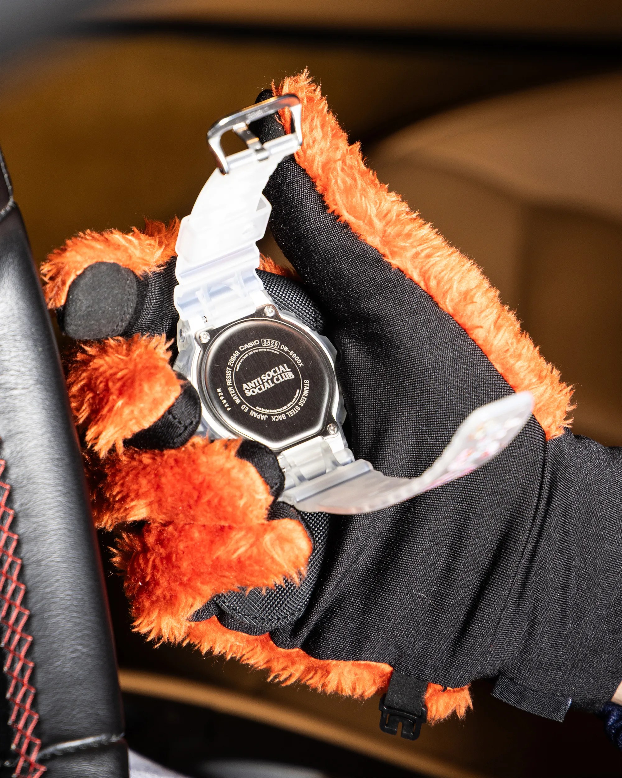 Hand wearing black and orange fuzzy gloves holding a clear Casio watch with "Anti Social Social Club" text on the back.